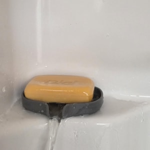 Könnte beinhalten: Ein gelbes Seifenstück liegt in einer grauen Seifenschale mit einem Ausguss. Die Seifenschale ist auf einer weißen Oberfläche montiert, und Wasser fließt aus dem Ausguss.