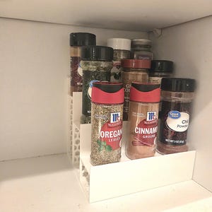 May include: A white tiered spice rack filled with various spice jars. Jars of oregano leaves, cinnamon ground, and chili powder are visible. The McCormick brand is featured on several spice containers.