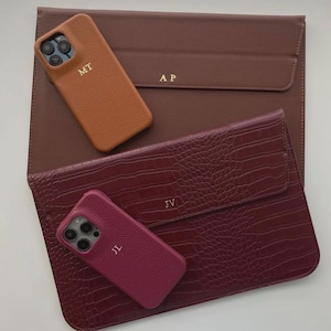 Può includere: Un assortimento di custodie per telefono e custodie per tablet in pelle personalizzate. Le custodie per telefono sono color cuoio e magenta, con le iniziali "MT" e "JL". Le custodie per tablet sono marroni e bordeaux, con le iniziali "AP" e "JV".