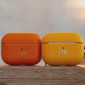 Peut inclure: Deux étuis AirPod personnalisés. L'un est orange avec "LBS" en lettres dorées, l'autre est jaune avec "MK" en lettres argentées. Les deux étuis ont un aspect texturé semblable au cuir.