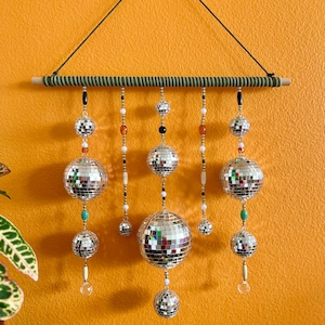 May include: A decorative wall hanging featuring multiple mirrored disco balls of varying sizes. The disco balls are suspended from a wooden dowel wrapped in green fabric. Strands of beads in various colors and shapes hang between the disco balls.