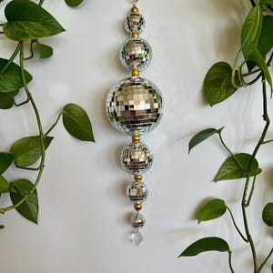 May include: A decorative hanging ornament featuring multiple silver mirrored disco balls of varying sizes, connected by gold-colored beads and a clear crystal at the bottom. The ornament is suspended from a small gold ring. Green leafy vines frame the ornament.