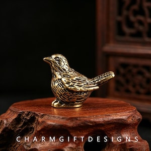 Puede incluir: Una pequeña figura de pájaro de latón macizo con líneas grabadas detalladas que representan las plumas en las alas, el cuerpo y la cola. El metal tiene un acabado pulido de color marrón dorado con áreas hundidas más oscuras.
