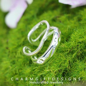 Puede incluir: Un anillo plateado brillante con un diseño único y fluido. El anillo tiene una forma curva y abierta con pequeños recortes circulares. El anillo está sobre un fondo de musgo verde vibrante. El texto "CHARMGIFTD ESIGNS Handcrafted Jewellery" está en la parte inferior.