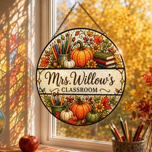 May include: A round stained glass suncatcher featuring autumn-themed illustrations of pumpkins, fall leaves, and art supplies. The centre text reads Mrs. Willow's CLASSROOM. The piece hangs from a metal chain and suction cup against a window.