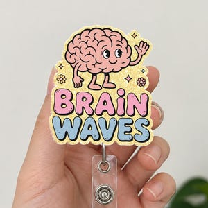 May include: A badge reel with a cartoon brain character and the words "BRAIN WAVES" in pink and blue. The brain is pink with black eyes and is waving. The reel has a clear plastic holder and a silver metal clip.