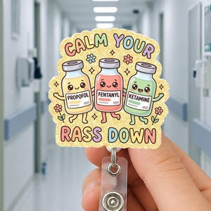 May include: A badge reel with the text "CALM YOUR RASS DOWN" and illustrations of three animated medicine bottles labeled "PROPOFOL", "FENTANYL", and "KETAMINE". The badge reel is held in a hand.