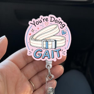 Puede incluir: Un portacredenciales circular con fondo rosa claro, una ilustración de un cinturón de transferencia blanco y el texto "You're Doing GAIT" en letras azules y negras. El diseño incluye pequeños corazones azules y estrellas rosas sobre una base blanca.