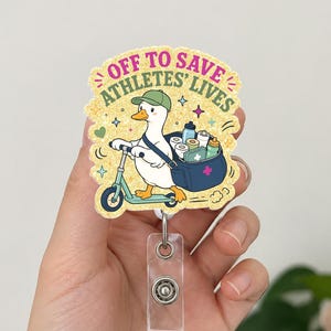 May include: A badge reel featuring a cartoon duck riding a scooter, carrying medical supplies. The text "OFF TO SAVE ATHLETES' LIVES" is displayed in a yellow and green font. The reel has a clear plastic holder and a metal clip.