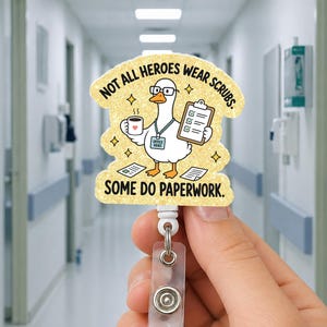 May include: A glittery badge reel featuring a cartoon goose wearing glasses and an ID badge that reads OFFICE HERO. The goose holds a coffee mug and a clipboard. The text reads NOT ALL HEROES WEAR SCRUBS. SOME DO PAPERWORK. The design is yellow and gold.
