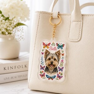 May include: A rectangular acrylic keychain with scalloped edges featuring a portrait of a Yorkshire Terrier, the name Belle in pink script, and colorful butterfly and floral illustrations, attached to a gold-toned metal ring and chain.