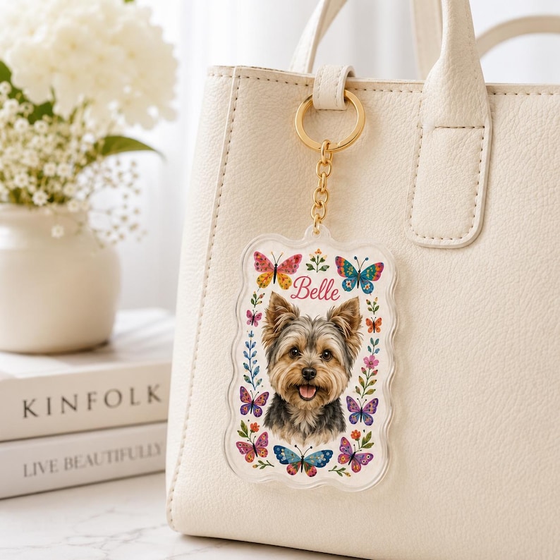 May include: A rectangular acrylic keychain with scalloped edges featuring a portrait of a Yorkshire Terrier, the name Belle in pink script, and colorful butterfly and floral illustrations, attached to a gold-toned metal ring and chain.