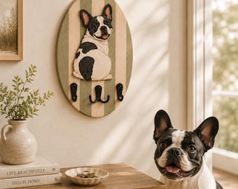 May include: An oval wall-mounted hook rack featuring a vertical striped pattern in light green and natural wood tones. A sculpted black and white French Bulldog figure is attached to the center. It includes three black metal hooks for hanging items.