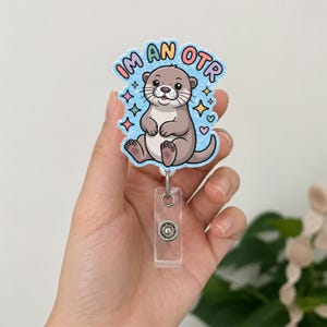 May include: A badge reel featuring a cartoon otter with the text "IM AN OTR" in a playful font. The otter is light brown with a white belly, surrounded by blue sparkles and colorful stars and hearts. The reel has a clear plastic holder.
