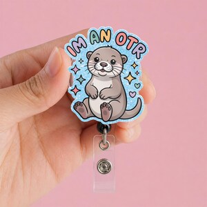 May include: A badge reel with a cartoon otter illustration. The badge reel has a light blue background with the words "I'm an OTR" in a colorful bubble font. The otter is gray and white with a smiling face and is surrounded by colorful stars and hearts.