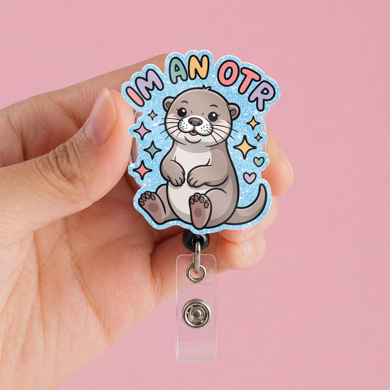 May include: A badge reel with a cartoon otter illustration. The badge reel has a light blue background with the words "I'm an OTR" in a colorful bubble font. The otter is gray and white with a smiling face and is surrounded by colorful stars and hearts.
