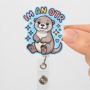 May include: A badge reel with a cartoon otter illustration. The otter is gray and white, with the text "I'M AN OTR" in a colorful bubble design. The badge reel has a clear plastic base and a metal clip.