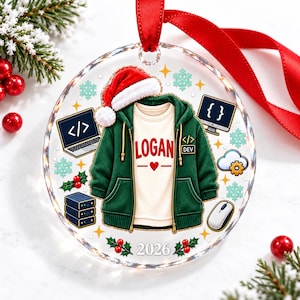 May include: A round clear acrylic Christmas ornament featuring a green zip-up hoodie over a white shirt with the name LOGAN in red text. The design includes a Santa hat, a laptop with code symbols, a computer monitor, a mouse, and the year 2026.