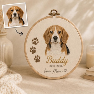 Puede incluir: Un bastidor de bordado circular de madera con un retrato bordado detallado de un perro beagle. La tela muestra tres huellas de patas marrones, el nombre Buddy, los años 2011-2026 y el texto Love, Mom con un pequeño símbolo de corazón.