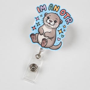 May include: A badge reel with a cartoon otter illustration. The otter is light brown with a white belly and is surrounded by a blue glittery border with the text "I'M AN OTR". The reel has a clear plastic strap and a metal snap.