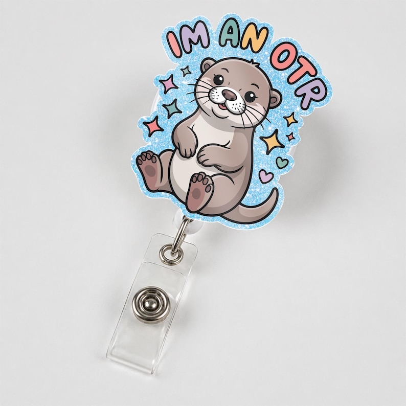 May include: A badge reel with a cartoon otter illustration. The otter is light brown with a white belly and is surrounded by a blue glittery border with the text "I'M AN OTR". The reel has a clear plastic strap and a metal snap.
