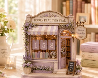 May include: A miniature bookshop model titled Violet's Bookshop with a lavender and white color scheme. The facade features the text BOOKS READ THIS YEAR, a hanging sign, a small chalkboard reading One More Chapter, and floral accents.