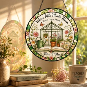 May include: A circular stained glass suncatcher featuring a greenhouse illustration, a watering can, an open book, and flowers. The text reads: Where Little Minds Bloom, Grow. Learn. Shine. Mrs. Miller. The border has a green and pink floral pattern.