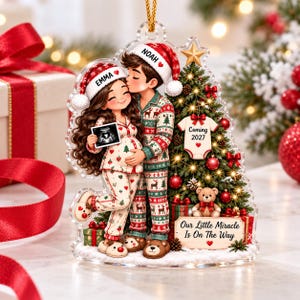 May include: A clear acrylic Christmas ornament featuring a couple in pajamas, with the names Emma and Noah on their hats. The woman holds an ultrasound image. A decorated tree displays a baby onesie labeled Coming 2027 and a sign reading Our Little Miracle Is On The Way.