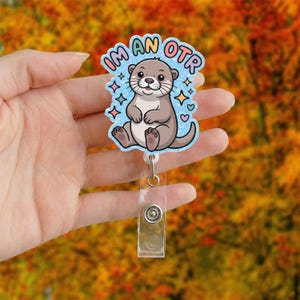 May include: A badge reel with a cartoon otter design. The otter is light brown with a white belly, sitting on a light blue background with the words "IM AN OTR". The badge reel has a clear plastic clip and a metal button.