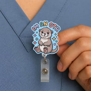 May include: A badge reel featuring a cartoon otter with the text "I'M AN OTR" in a blue glitter design. The otter is gray and brown, surrounded by stars and hearts. The reel has a clear plastic holder and a metal button.