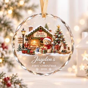 May include: A round, clear glass Christmas ornament with a faceted edge and a gold ribbon. The design features a snowy Santa's Toy Shop, a teddy bear, a toy train, a nutcracker, and a Christmas tree. Text reads: Jayden's FIRST CHRISTMAS 2026.