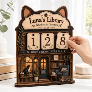 May include: A wooden cat-shaped calendar titled Luna's Library Whiskers & Chapters 2026. It features three interchangeable number tiles, the text BOOKS READ THIS YEAR, a miniature library scene with two cats, a blue armchair, and a door with an OPEN sign.