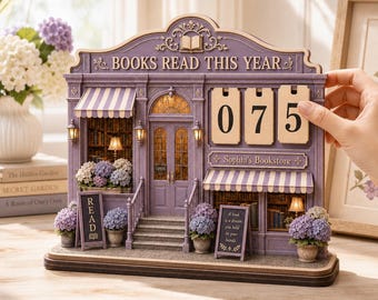 May include: A purple miniature bookstore tracker with the text BOOKS READ THIS YEAR and Sophia's Bookstore. It features three wooden number tiles showing 075, striped awnings, miniature bookshelves, and potted hydrangea flowers on a base.