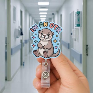 May include: A badge reel featuring a cartoon otter with the text "I'M AN OTR". The otter is light brown with a blue background and colorful star and heart accents. The reel has a clear plastic clip and a metal snap.