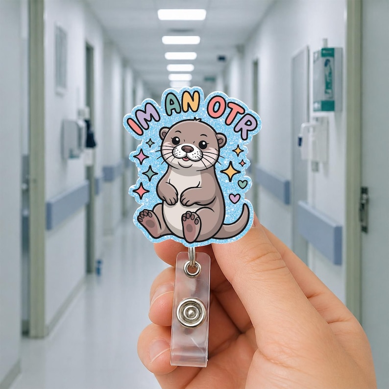 May include: A badge reel featuring a cartoon otter with the text "I'M AN OTR". The otter is light brown with a blue background and colorful star and heart accents. The reel has a clear plastic clip and a metal snap.