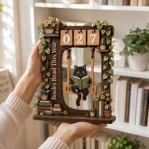 May include: A dark wood perpetual calendar featuring a black cat sitting on a swing and reading a book titled Sophia. The frame is decorated with vines, potted plants, and stacks of books, with the text Books Read This Year and the numbers 027.