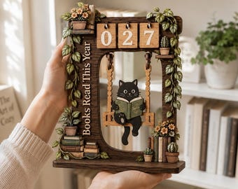 May include: A dark wood perpetual calendar featuring a black cat sitting on a swing and reading a book titled Sophia. The frame is decorated with vines, potted plants, and stacks of books, with the text Books Read This Year and the numbers 027.