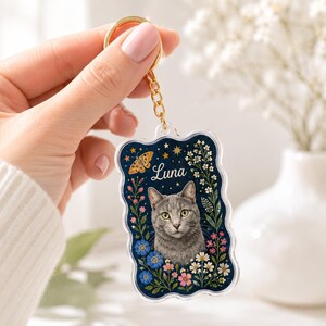 May include: A rectangular acrylic keychain with scalloped edges featuring a portrait of a gray cat surrounded by colorful flowers, a moth, and stars on a dark blue background. The name Luna is printed in white script above the cat's head.