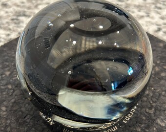Puede incluir: Una esfera de vidrio transparente con un interior oscuro y arremolinado y pequeñas burbujas de aire. La esfera descansa sobre una superficie oscura y texturizada. La esfera refleja la luz y el entorno. El texto "moodmats.com" y "scraps" es visible.