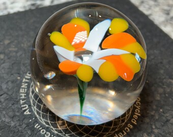 Puede incluir: Un pisapapeles esférico de cristal transparente con un diseño floral colorido. La flor presenta pétalos naranjas, amarillos y blancos y un tallo verde. Se encuentra sobre una superficie gris oscuro.