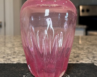 Puede incluir: Un jarrón de cristal rosa con un diseño texturizado y acanalado. El jarrón tiene una parte superior redondeada y una base ligeramente ensanchada. El color es un degradado, con un rosa más oscuro en la base y un rosa más claro en la parte superior. El jarrón está sobre una superficie negra.