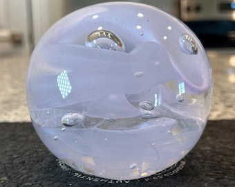 Puede incluir: Un pisapapeles esférico de vidrio transparente. El interior presenta patrones arremolinados blancos y lavanda, con varias burbujas de aire. El pisapapeles está sobre una superficie oscura.