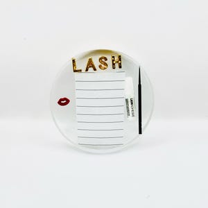 Könnte beinhalten: Runde, transparente Wimpernstation mit goldenen Glitzerbuchstaben mit der Aufschrift "LASH". Ein kleiner Notizblock, eine rote Lippen-Grafik und eine Tube Wimpernkleber sind ebenfalls sichtbar. Zum Auftragen von falschen Wimpern.