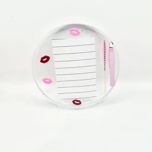 Könnte beinhalten: Ein klarer, runder Harztablett mit einem linierten Notizblock und rosa Wimpernwerkzeugen. Das Tablett ist mit rosa und weinroten Lippenmotiven verziert. Eine rosa Wimpernbürste und eine Pinzette sind sichtbar.