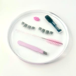 Könnte beinhalten: Eine transparente, runde Ablage enthält falsche Wimpern, Wimpernkleber, einen rosa Lippen-Charm, eine Wimpernbürste und eine rosa Pinzette. Die Wimpernkleber-Tube ist schwarz mit weißem Text. Auf weißem Hintergrund angeordnet.