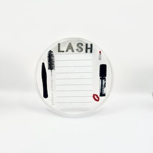 Könnte beinhalten: Runde, transparente Wimpernstation mit dem Wort "LASH" in silbernen Buchstaben. Die Station enthält eine schwarze Mascarabürste, eine Pinzette, Lippenstift und eine Flasche Wimpernkleber. Die Station hat einen linierten Bereich für Notizen.