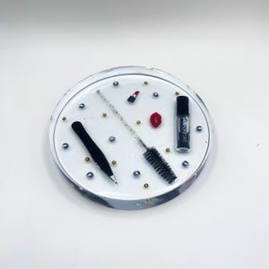 Könnte beinhalten: Ein klarer, runder Harz-Tablett mit eingebetteten silbernen und goldenen Perlen. Das Tablett enthält Miniatur-Make-up-Artikel: eine schwarze Pinzette, eine Mascarabürste, einen Lippenstift, eine Lippe und eine Tube Wimpernkleber. Der Text "LASH ADHESIVE" ist sichtbar.