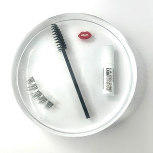 Könnte beinhalten: Ein klarer, kreisförmiger Behälter enthält eine schwarze Mascarabürste, ein Paar falsche Wimpern, einen kleinen roten Lippen-förmigen Anhänger und eine weiße Flasche L.A. Colors Wimpernkleber. Die Artikel sind auf weißem Hintergrund angeordnet.