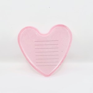Könnte beinhalten: Ein rosa herzförmiger Notizblock mit liniertem Papier. Der Notizblock besteht aus einem durchscheinenden Material und hat eine glatte Oberfläche. Die Linien sind schwarz und gleichmäßig angeordnet. Die Herzform ist weich und abgerundet.