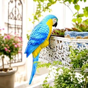 May include: A decorative parrot figurine featuring vibrant blue and yellow feathers, a white face patch, and a black beak. The bird is designed with a curved metal hook attached to its underside, allowing it to hang securely from the edge of a planter.